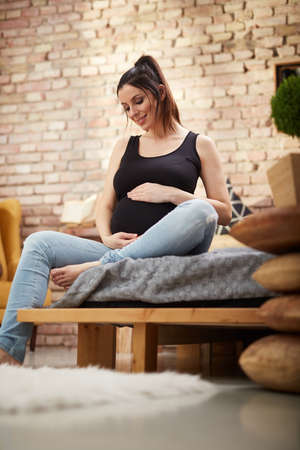 Happy pregnant woman sitting on bed at homeの写真素材