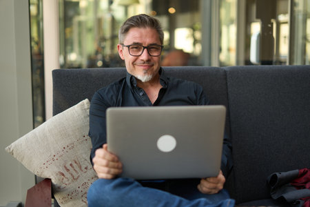 Business portrait - businessman sitting on sofa working with laptop computer in office, lobby or public space, Happy middle aged man smiling.の写真素材