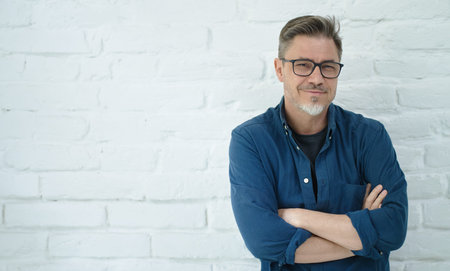 Portrait of happy casual middle aged man in glasses smiling. Copy space, white brick wall.