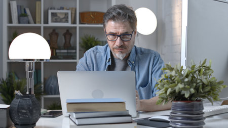 Older man working online with laptop computer at home sitting at desk. Home office, browsing internet, study room, entrepreneur.の写真素材