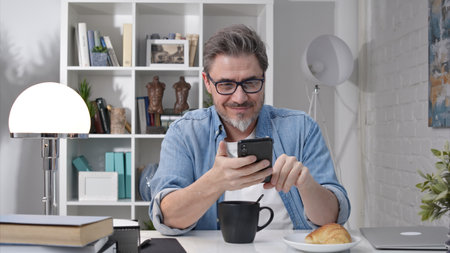 50s man having breakfast checking news on phone drinking morning coffee at home. Confident happy smiling.の写真素材