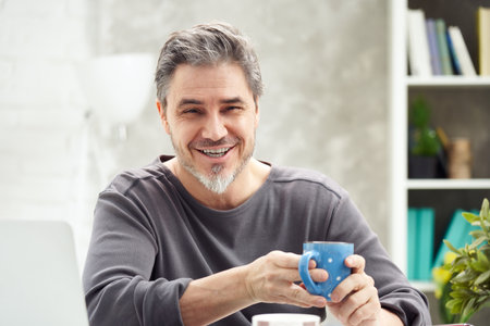 Portrait of happy man at home sitting at desk, drinking coffee, looking at camera. Happy smile, grey hair, beard. Portrait of mature age, middle age, mid adult man in 50s.の写真素材