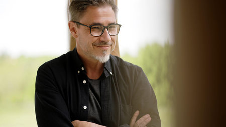 Portrait of happy casual middle age man in glasses standing on balcony at home relaxing, smiling.