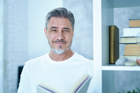 Portrait of mature age, middle age, mid adult casual man in glasses at home, happy smiling, gray hair, copy space. Standing at book self reading.の写真素材