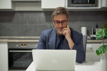 Businessman in home office working with laptop in kitchen. Man in mature age, middle age, bearded.の写真素材
