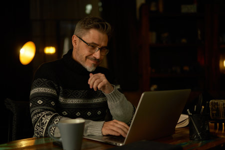 Older man sitting at desk in dark cosy room working on laptop computer in home office. Mature age, middle age, mid adult casual man in 50s, confident happy smiling.の写真素材