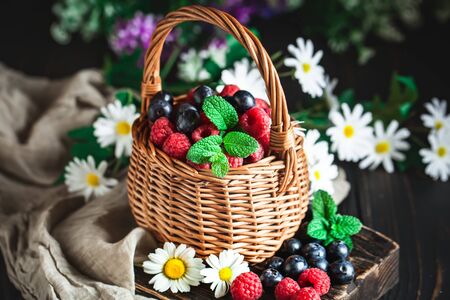 Raspberries and blueberries in a basket with chamomile and leaves on a dark background. Summer and healthy food concept. Selective focus.の写真素材