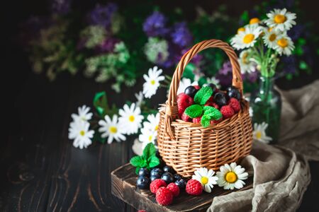 Raspberries and blueberries in a basket with chamomile and leaves on a dark background. Summer and healthy food concept. Background with copy space. Selective focus.の写真素材