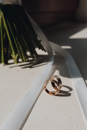 wo gold wedding rings rest on a sunlit white surface with a bouquet stem and satin ribbon, capturing...