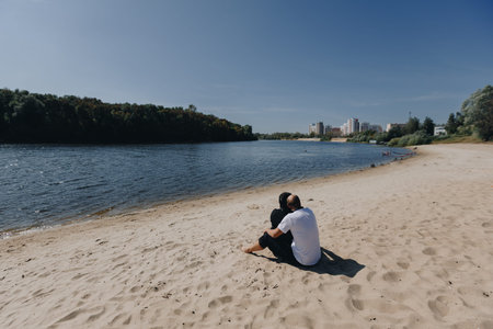 Romantic couple sits together on sandy beach, gazing at the river and distant city, enjoying tranquil summer day and quiet connection outdoorsの写真素材