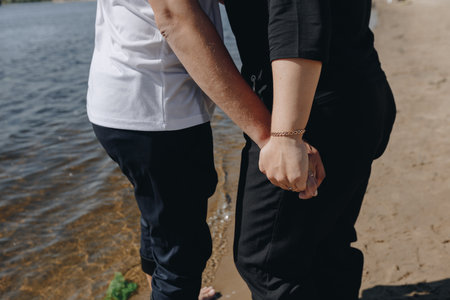 Close-up of a couple holding hands while standing on a sandy shore by the water, expressing love, connection, and romance in natureの写真素材