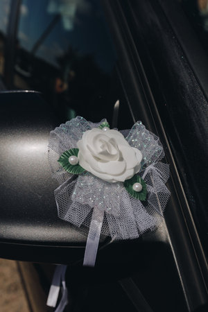 Elegant artificial white flower and lace bow decor attached to a dark car mirror, symbolizing stylish wedding transportation and festive celebration.の写真素材