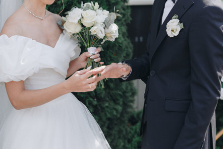 Bride in white dress and bouquet places wedding ring on the groom's hand during elegant outdoor ceremony, symbolizing unity and love.の写真素材