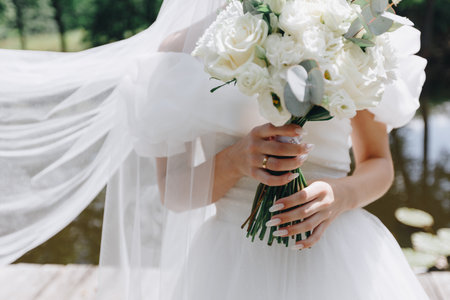 Bride in white dress holding bouquet of white roses and eucalyptus, with veil flowing in the wind near a pond, capturing romance and elegance of wedding dayの写真素材