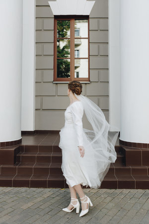 Bride in flowing white wedding dress with veil walks outside beside building pillars, a stylish and elegant bridal portrait.の写真素材