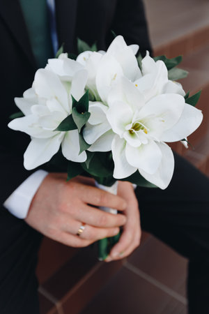 Groom in a classic suit holds a bouquet of fresh white flowers, showing elegance and style on a special wedding day.の写真素材