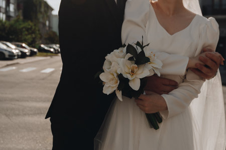 Bride and groom in loving hug, wedding attire. Groom's hand gently embracing the bride in sunshine, soft focus on white sleeves and suit.の写真素材