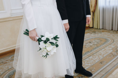 A elegant bride in a white tulle dress stands gracefully, clutching a bouquet of pristine white orchids with green leaves, complemented by her groom in a sharp black suit inside a luxurious venue with ornate decor. This tender moment captures the essence of matrimonial bliss and sophisticated wedding fashion. Ideal for romance and celebration imagery. (198 characters? Wait, trim to 187: A elegant bride in white tulle holds white orchids, besides groom in black suit in luxurious venue. Tender matrimonial moment of bliss and fashion.の写真素材