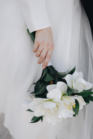 Bride's hand in white dress holds bouquet of white flowers, golden wedding band visible, detail from stylish wedding ceremony and celebration.の写真素材