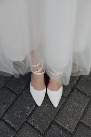 Artistic closeup of the bride's white pointed shoes peeking out from under her wedding dress, standing on stone pavement.の写真素材
