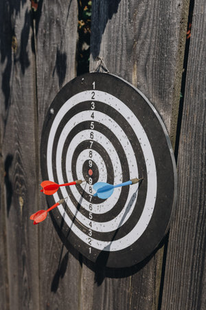 Classic black and white dartboard attached to an old wooden fence outdoors, hit by red and blue...