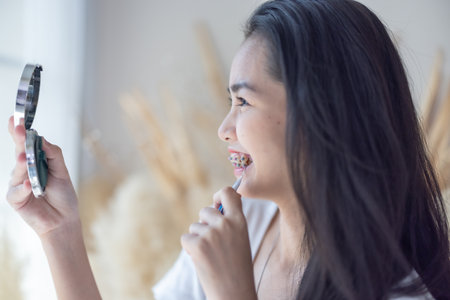 A young Asian girl with braces applies makeup, looking into a handheld mirror in a cozy, well-lit...