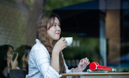 A contemplative Asian woman listens to music on red headphones while working on her laptop in a cozy...
