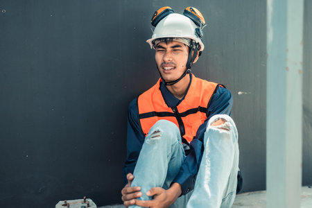 Asian HVAC engineer suffered serious leg injury on the job. Urgent first aid and assistance from coworkers is essential. Expressions of pain on face convey physical and emotional response.の写真素材