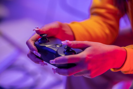 Close-up of female's hands with decorated nails holding gaming controller, illuminated by colorful...