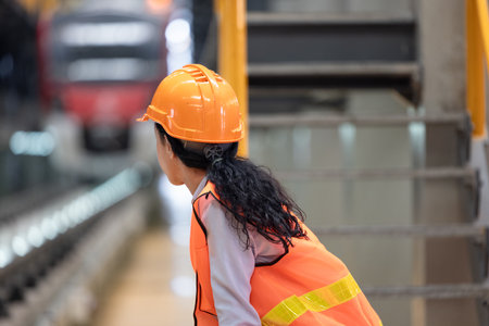 Female engineer with orange hard hat and safety vest is seen from behind, observing train inside maintenance depot. Highlights industrial work, safety, role of women in engineering and transportationの写真素材