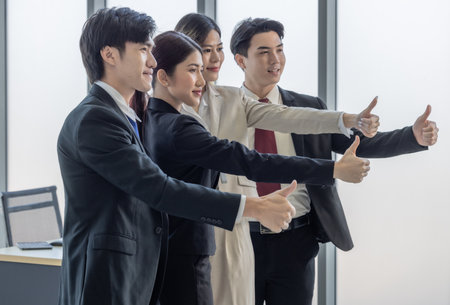 A group of four business professionals giving thumbs up in a modern office setting, symbolizing teamwork and success.の写真素材