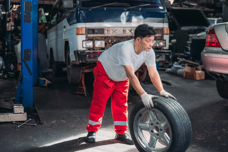 Mechanic in a workshop, rolling car tire towards a vehicle, showcasing the physical effort and nature of automotive repair work. The technician's dedication, strength in performing maintenance tasksの写真素材