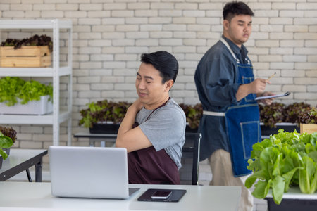 A weary gardener takes a break in urban farm, seated with laptop, showing signs of fatigue, shoulder pain. Fresh vegetables surround him, reflecting hard work and dedication involved in urban farmingの写真素材