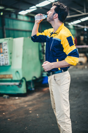 A factory worker in yellow and blue uniform takes refreshing drink from water bottle inside...