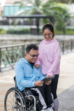 A cheerful husband in a wheelchair and his wife collaborate while using a tablet in an outdoor...