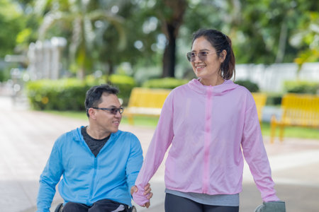 A couple enjoys sunny day at the park, with the man in wheelchair and the woman walking beside him....