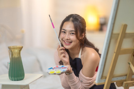 Young woman focuses intently on her artwork, painting on a canvas set up on an easel in her home studio. The scene captures a cozy, creative atmosphere with soft natural light filtering into the roomの写真素材