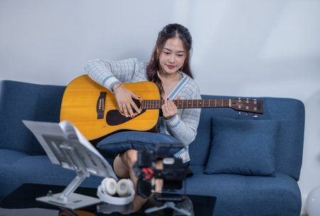 A young woman playing an acoustic guitar while smiling, sitting on a sofa with a music stand and...