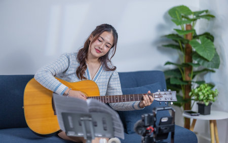 A young woman playing an acoustic guitar while smiling, sitting on a sofa with a music stand and recording equipment nearby, creating a cozy and creative atmosphere.の写真素材