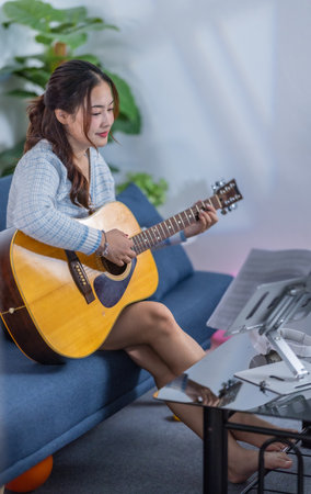 A young woman playing an acoustic guitar while smiling, sitting on a sofa with a music stand and recording equipment nearby, creating a cozy and creative atmosphere.の写真素材