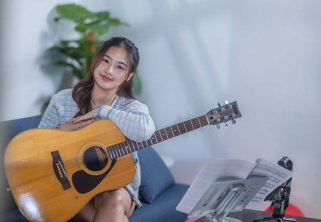 A young woman playing an acoustic guitar while smiling, sitting on a sofa with a music stand and recording equipment nearby, creating a cozy and creative atmosphere.の写真素材