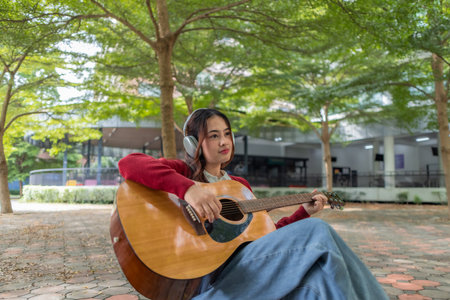 A young woman plays acoustic guitar in public space, immersed in music. Serene outdoor environment adds to calm and artistic vibe, creating a perfect backdrop for creative energy and performance.の写真素材