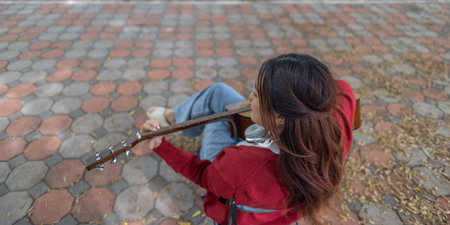 A young woman plays acoustic guitar in public space, immersed in music. Serene outdoor environment...