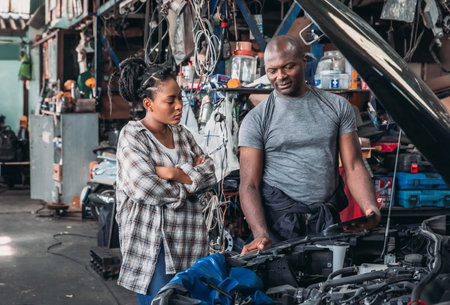 A woman watches intently as a mechanic discusses car issues under the hood of a vehicle in a cluttered workshop. Tools and parts are scattered throughout the space, signifying a busy workday.の写真素材