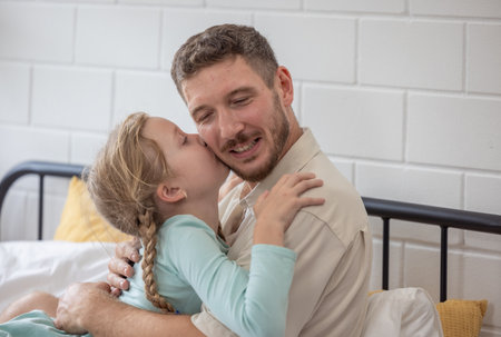 A man and his daughter are embracing on a bed covered with white sheets. The room is bright, and they both share smiles, creating a warm family atmosphere on a peaceful morning.の写真素材