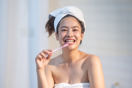 A woman smiles joyfully while brushing her teeth in a well-lit bathroom. She is wrapped in a towel and enjoys her morning routine after a refreshing shower.の写真素材