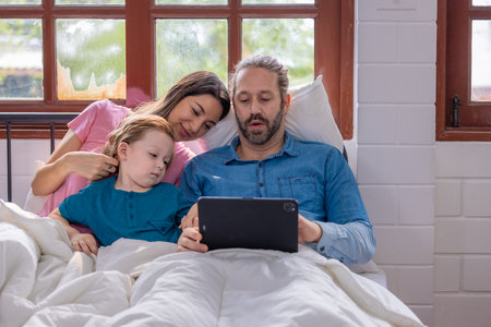 A mother leans on her partner while their child rests beside them in bed. They are engaged in watching content on a tablet, sharing a peaceful morning.の写真素材