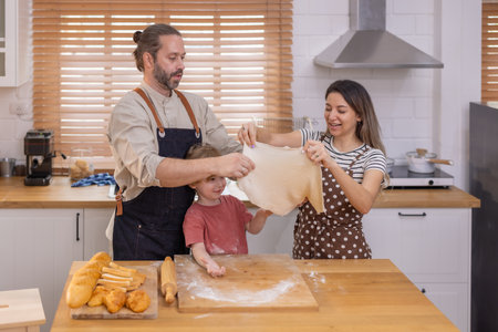 A family is happily engaged in baking at home, with a child playfully participating as they prepare dough on a wooden surface surrounded by bread.の写真素材