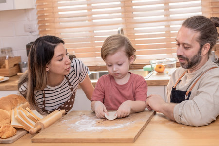 A family is happily engaged in baking at home, with a child playfully participating as they prepare dough on a wooden surface surrounded by bread.の写真素材