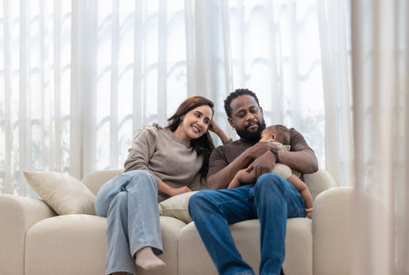 A loving black family sitting together on a cozy sofa in a bright living room. The father gently holds a baby while the mother smiles warmly, creating a scene of comfort and togetherness.の写真素材
